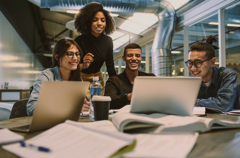 Werkstudierende Gruppe von vier jungen Menschen arbeitet gemeinsam an einem Laptop in einem hellen Arbeitsraum.
