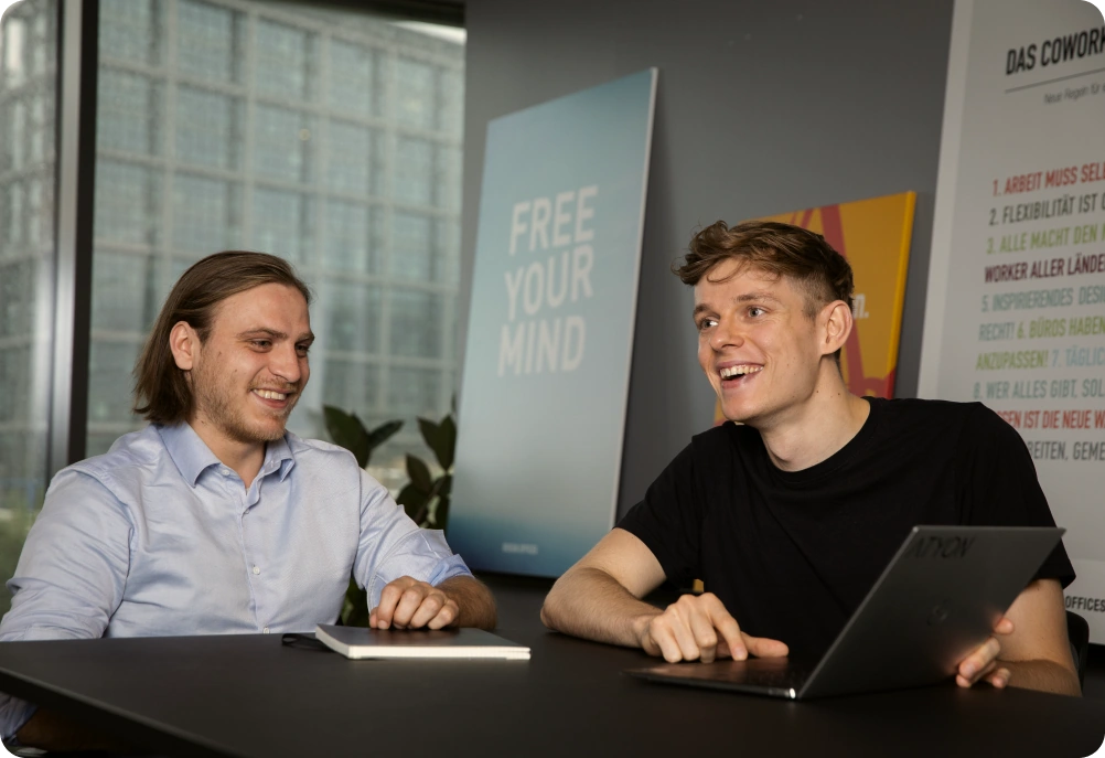 Zwei junge Männer sitzen im Büro am Tisch und lachen gemeinsam, vor ihnen liegen ein Laptop und ein Notizbuch.