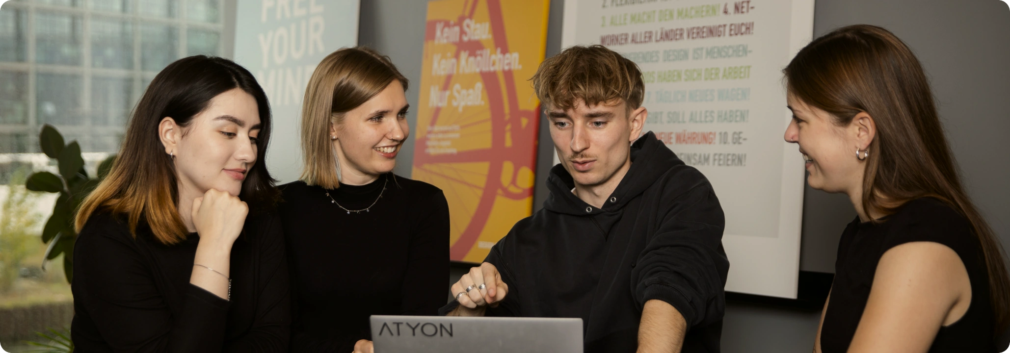 Vier junge Menschen, drei Frauen und ein Mann, schauen auf den Laptop-Bildschirm in einem Büroraum und sprechen miteinander, während sie lächeln.