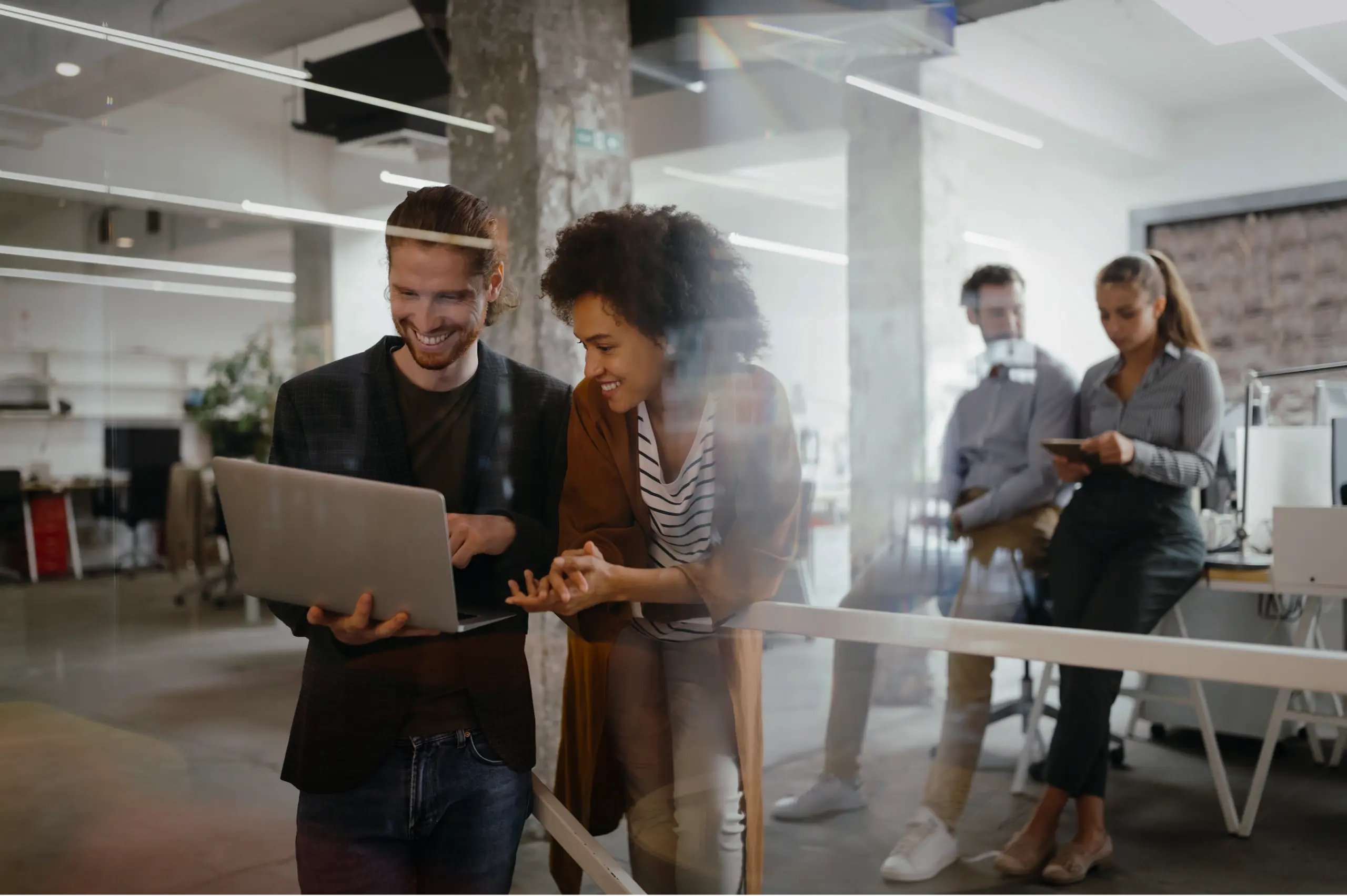 Teamgespräch im Büro Ein Mann und eine Frau stehen lächelnd im Vordergrund und schauen auf ein Laptop, während im Hintergrund weitere Personen, ein Mann und eine Frau, miteinander sprechen.
