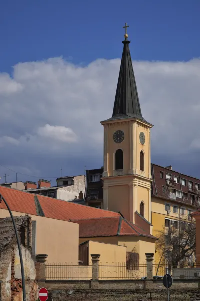 Die Kirche in Pančevo, ein markantes Gebäude mit einem hohen, schlanken Glockenturm, gekrönt von einem Kreuz. Der Turm verfügt über eine sichtbare Uhr. Neben der Kirche sind verschiedene städtische Gebäude mit roten und gelben Fassaden zu sehen, alles unter einem dramatischen Himmel mit Wolken.