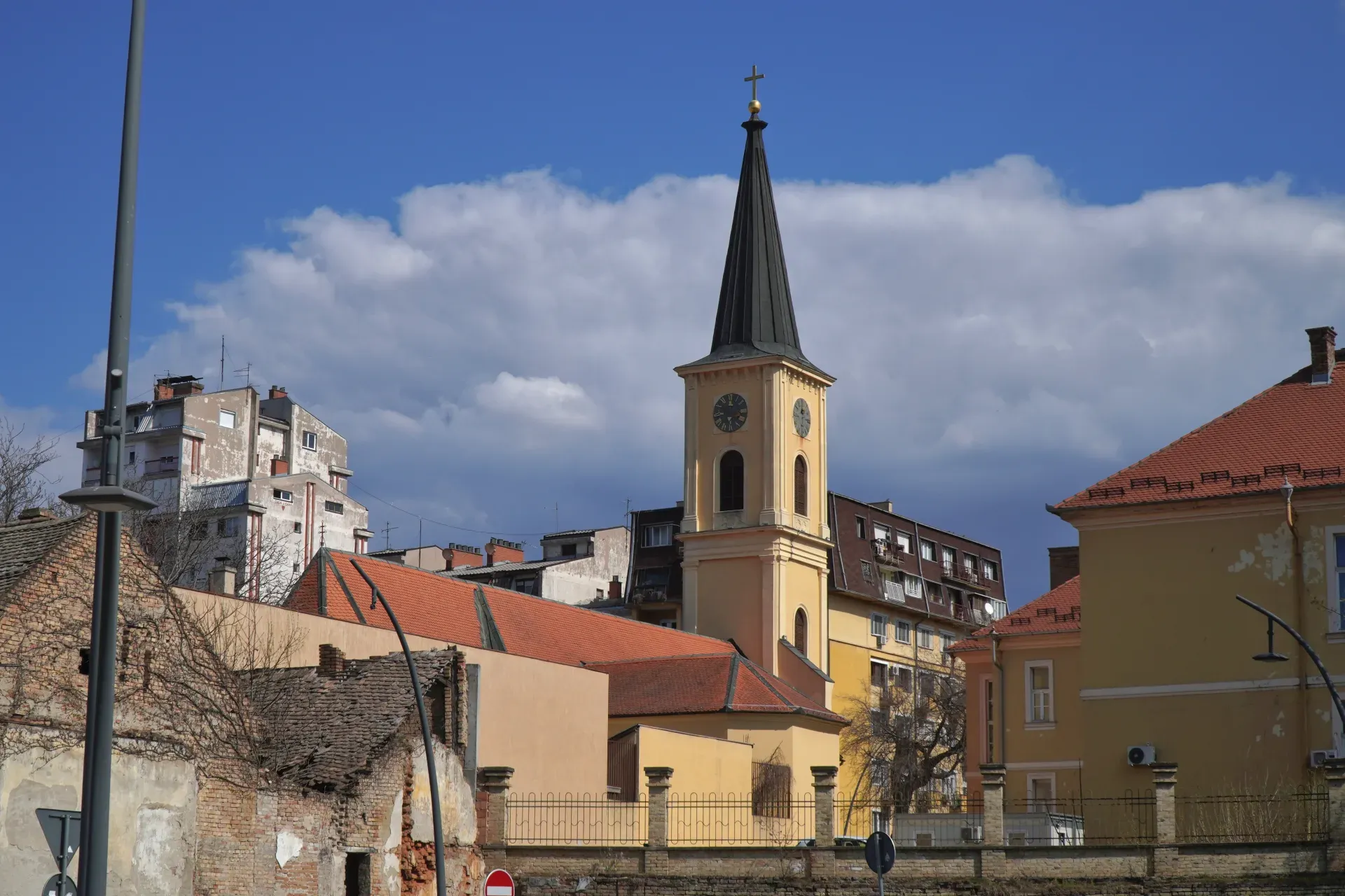 Ein gelber Kirchturm mit einem schwarzen spitzen Dach und einer Uhr ist in Pančevo, Serbien, zu sehen. Moderne Gebäude sind im Hintergrund zu sehen, und der Himmel ist blau mit weißen Wolken.