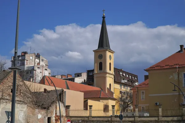 Ein gelber Kirchturm mit einem schwarzen spitzen Dach und einer Uhr ist in Pančevo, Serbien, zu sehen. Moderne Gebäude sind im Hintergrund zu sehen, und der Himmel ist blau mit weißen Wolken.