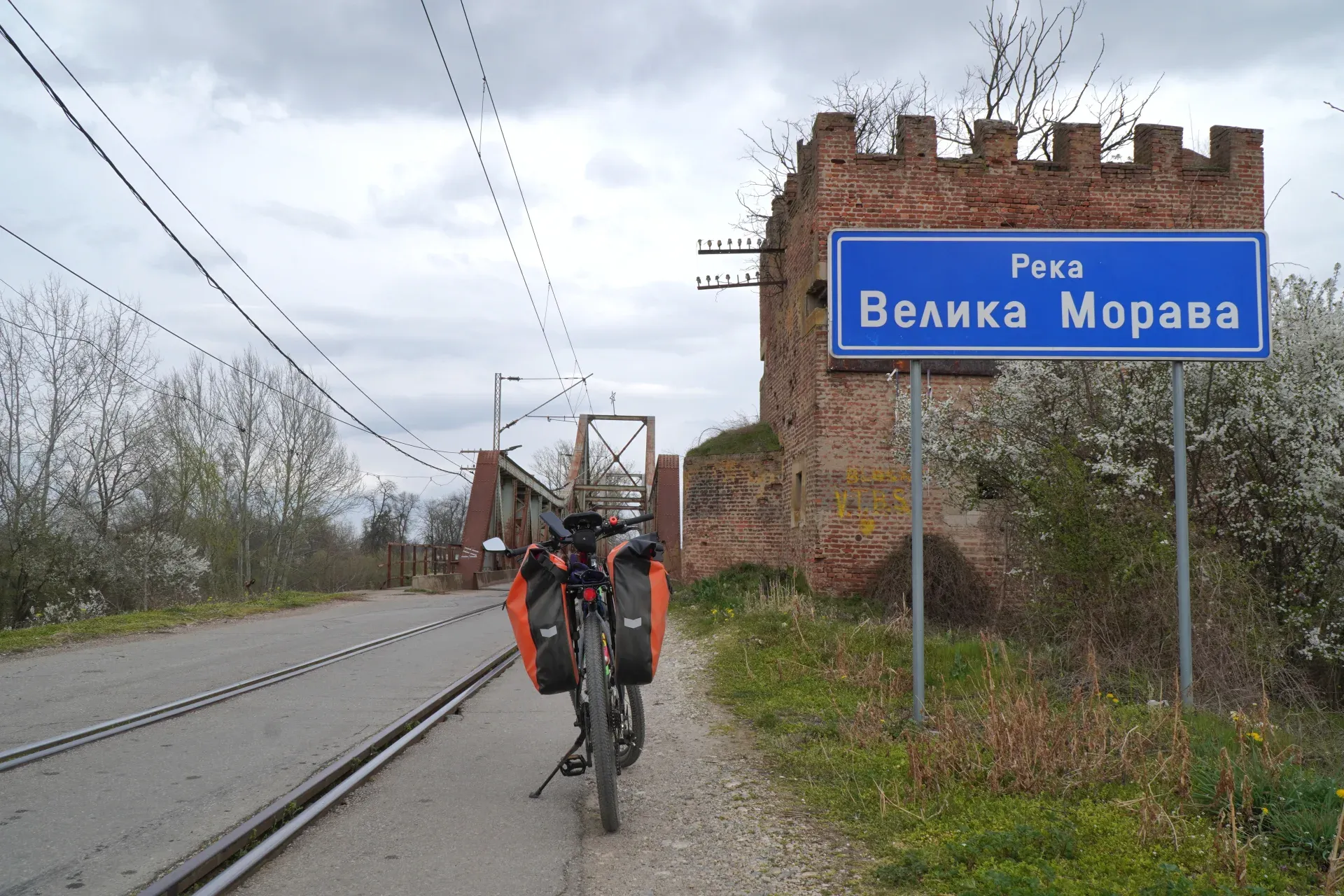 Ein mit orangefarbenen Satteltaschen beladenes Fahrrad steht auf einem Schotterweg neben Bahngleisen. Im Hintergrund ist der Ljubičevski Most (Brücke) über die Große Morava zu sehen, zusammen mit einem alten Backsteinturm und einem blauen Schild, auf dem 'Velika Morava' steht. Die Szene befindet sich in der Nähe von Požarevac, Serbien.