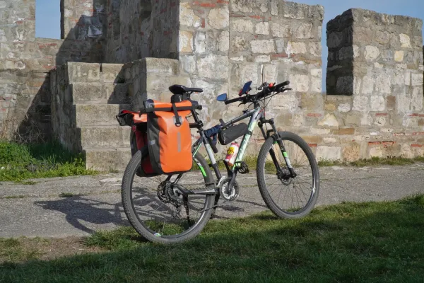 Una bicicleta cargada con alforjas naranjas se encuentra en un camino pavimentado frente a una antigua muralla de piedra de la Fortaleza de Smederevo, Serbia.