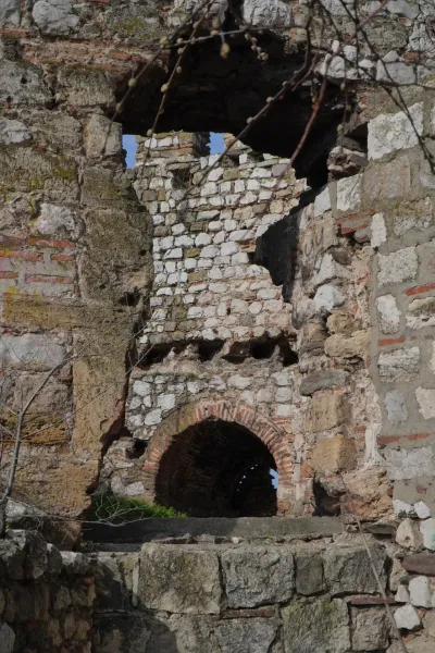 Un antiguo complejo de piedra con un pasaje redondeado y agujeros en las paredes, a través de los cuales se puede ver el cielo azul. Algunas ramas con capullos sobresalen desde arriba en la imagen.