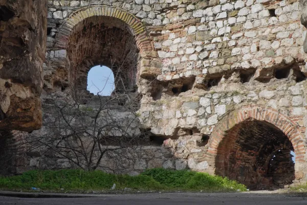 Una pared de piedra en ruinas con varios arcos y aberturas a través de las cuales se ve el cielo azul, así como ramas secas y vegetación verde en el suelo.
