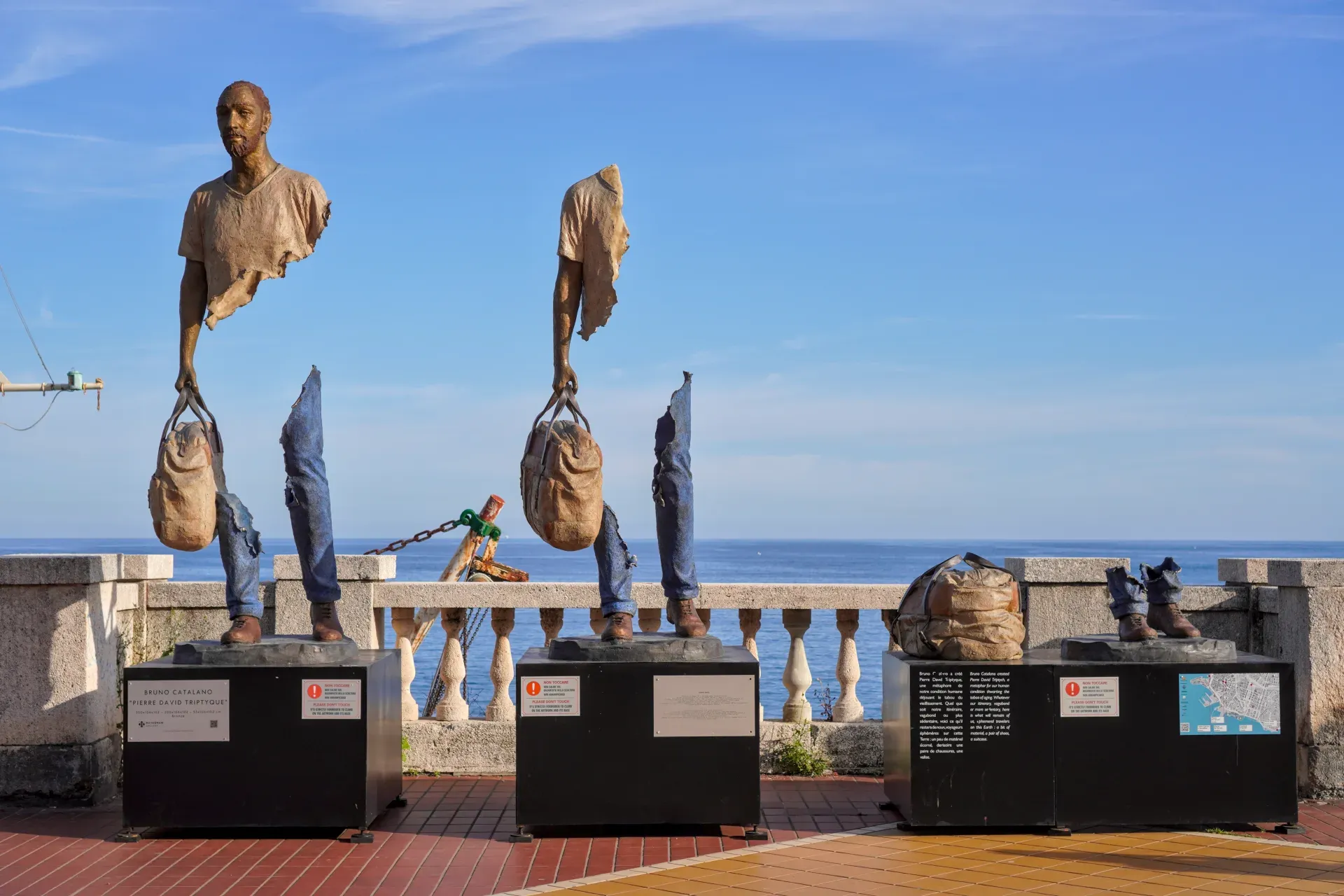 Esculturas fragmentadas de bronce de torsos y piernas humanas portando bolsas, expuestas sobre pedestales a lo largo del paseo marítimo de Génova con el mar azul y el cielo al fondo.