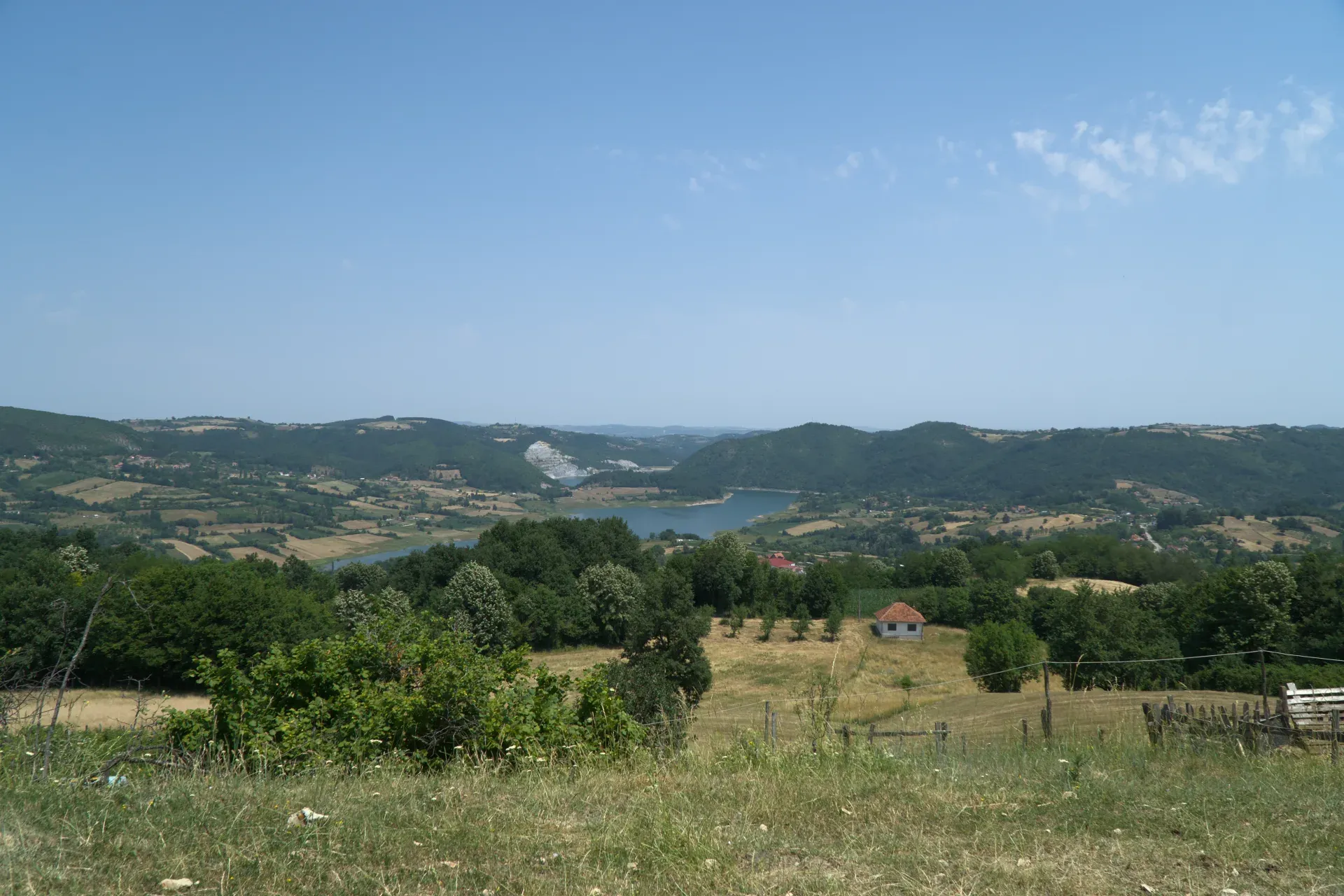 Brdoviti pejzaž sa jezerom i zelenim drvećem, na putu za Nacionalni park Tara u Srbiji, sa malom kućom u daljini.