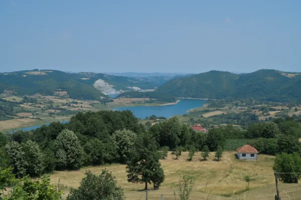 Un vaste paysage avec des collines vertes et boisées, des champs cultivés et un lac turquoise dans le parc national de Tara, Serbie. Une petite maison blanche au toit rouge se dresse dans un champ entouré d'arbres. À l'arrière-plan, d'autres collines et le vaste ciel sont visibles.