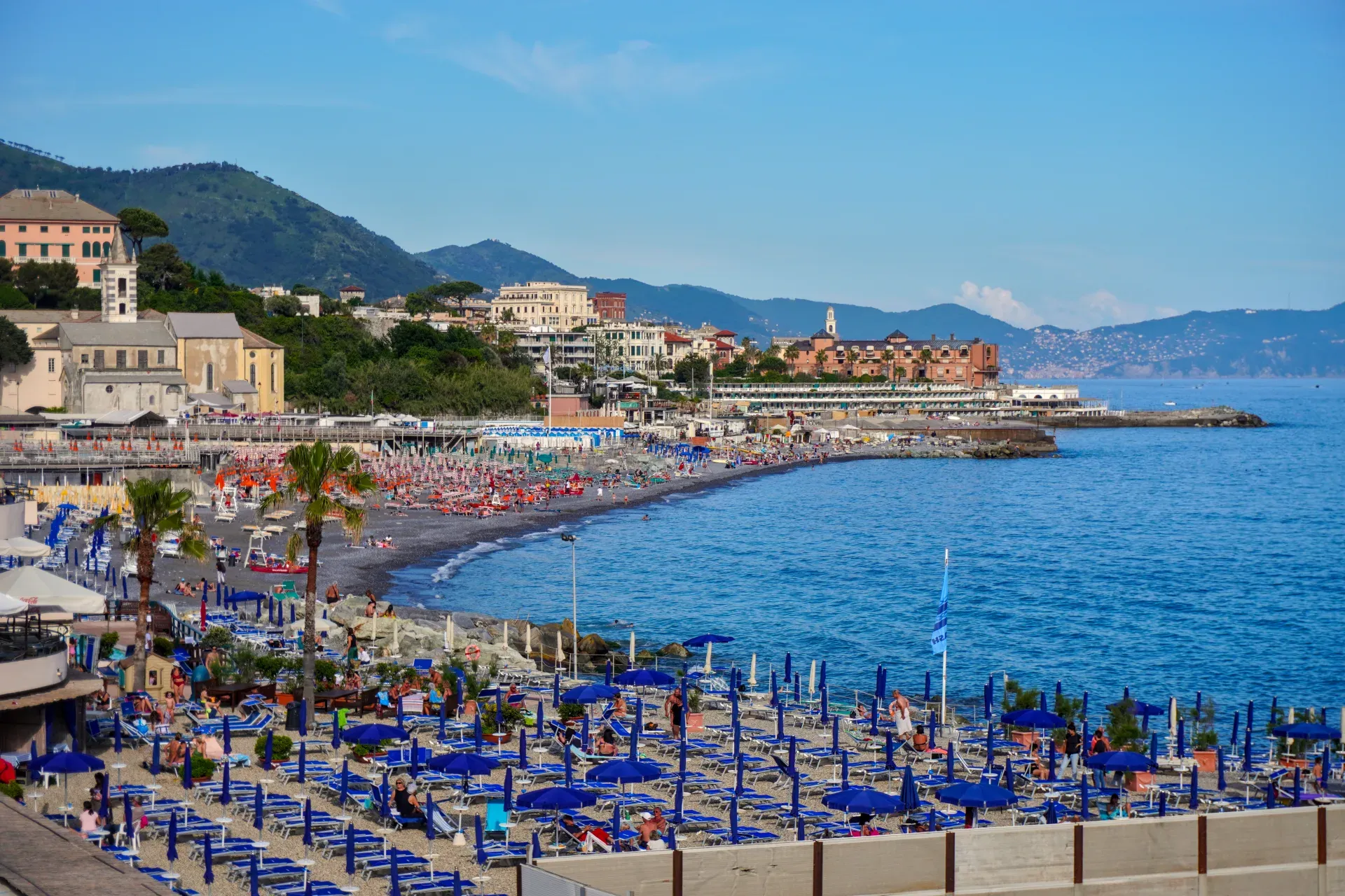 Una spiaggia affollata a Genova, Italia, con numerosi ombrelloni e lettini blu, edifici e colline verdi sullo sfondo, e il mare blu sulla destra.