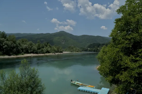 Širok pogled na reku Drinu sa zelenim, šumovitim brdima u pozadini. Mali čamac je usidren na obali sa plutajućim dokom pored njega. Tirkizna voda je mirna i odražava plavo nebo sa retkim oblacima.