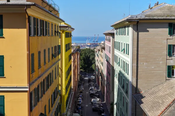 A colorful Italian alley with pastel-colored buildings, cars, and a view of the sea and harbor cranes.