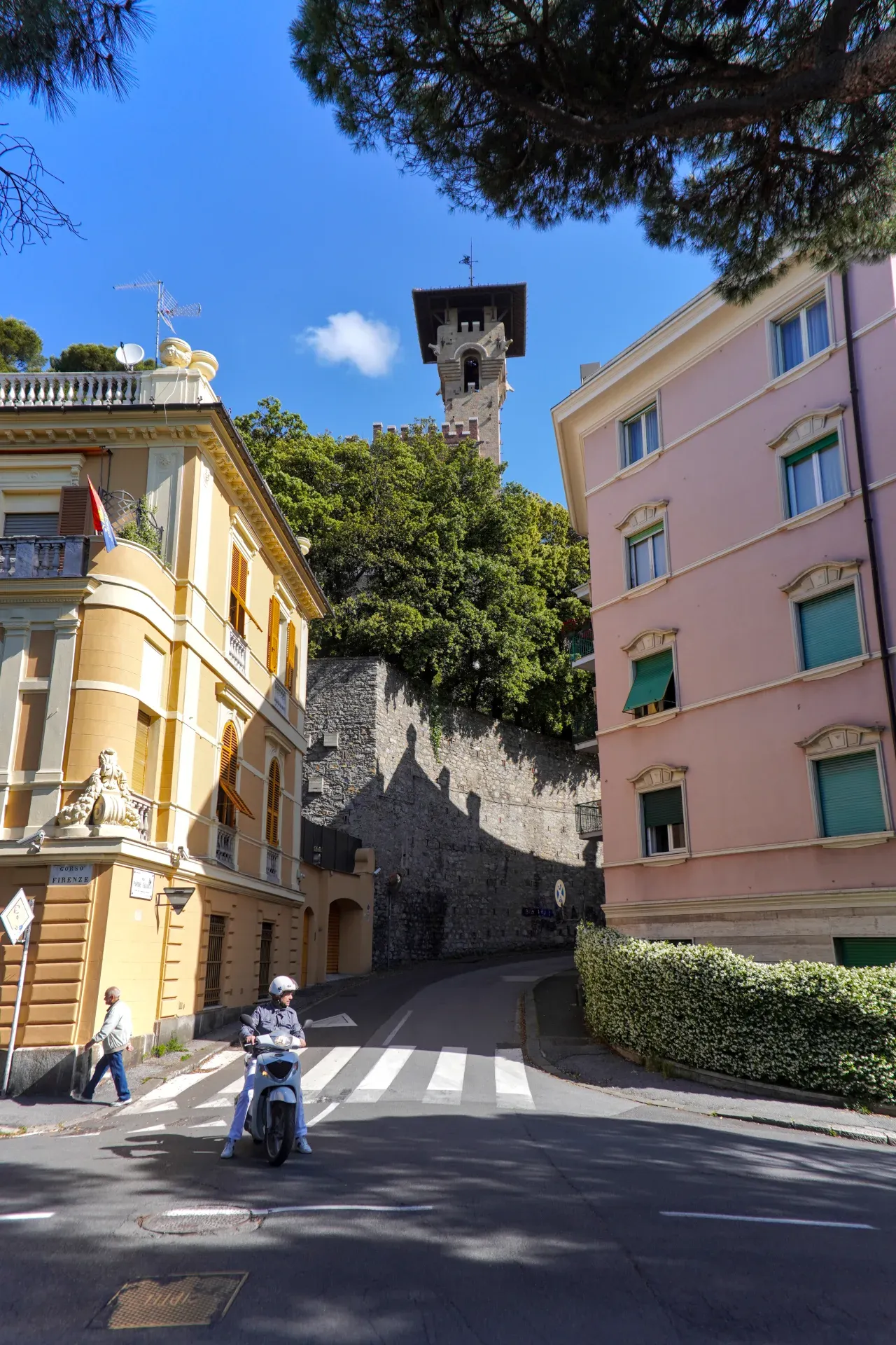 Una llamativa torre italiana se eleva sobre árboles y edificios pastel en Génova, con una moto en una calle al frente.