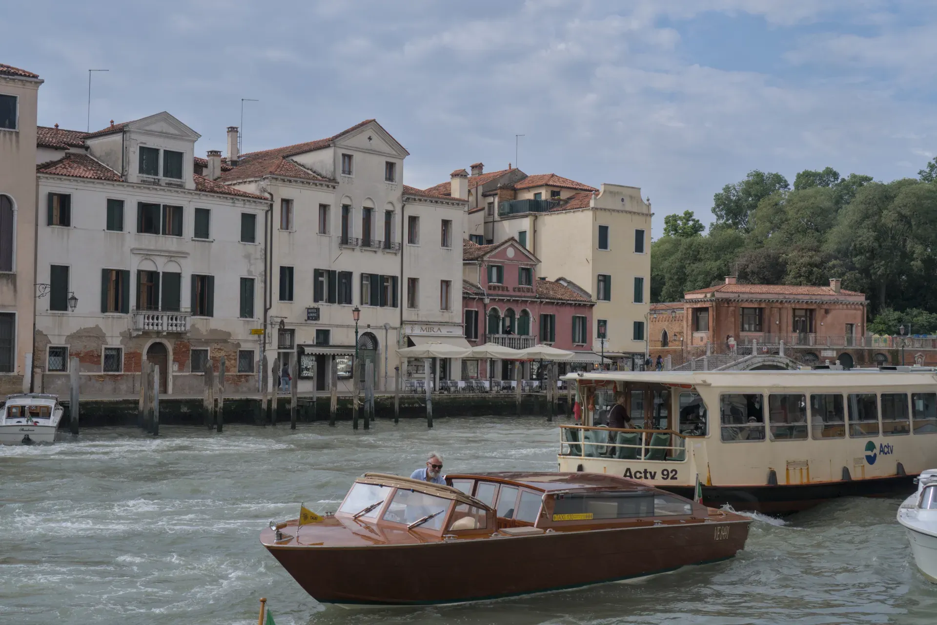 Un canal vénitien avec la vue de face d'un taxi nautique marron et un navire de passagers plus grand, entouré de bâtiments classiques sous un ciel nuageux.