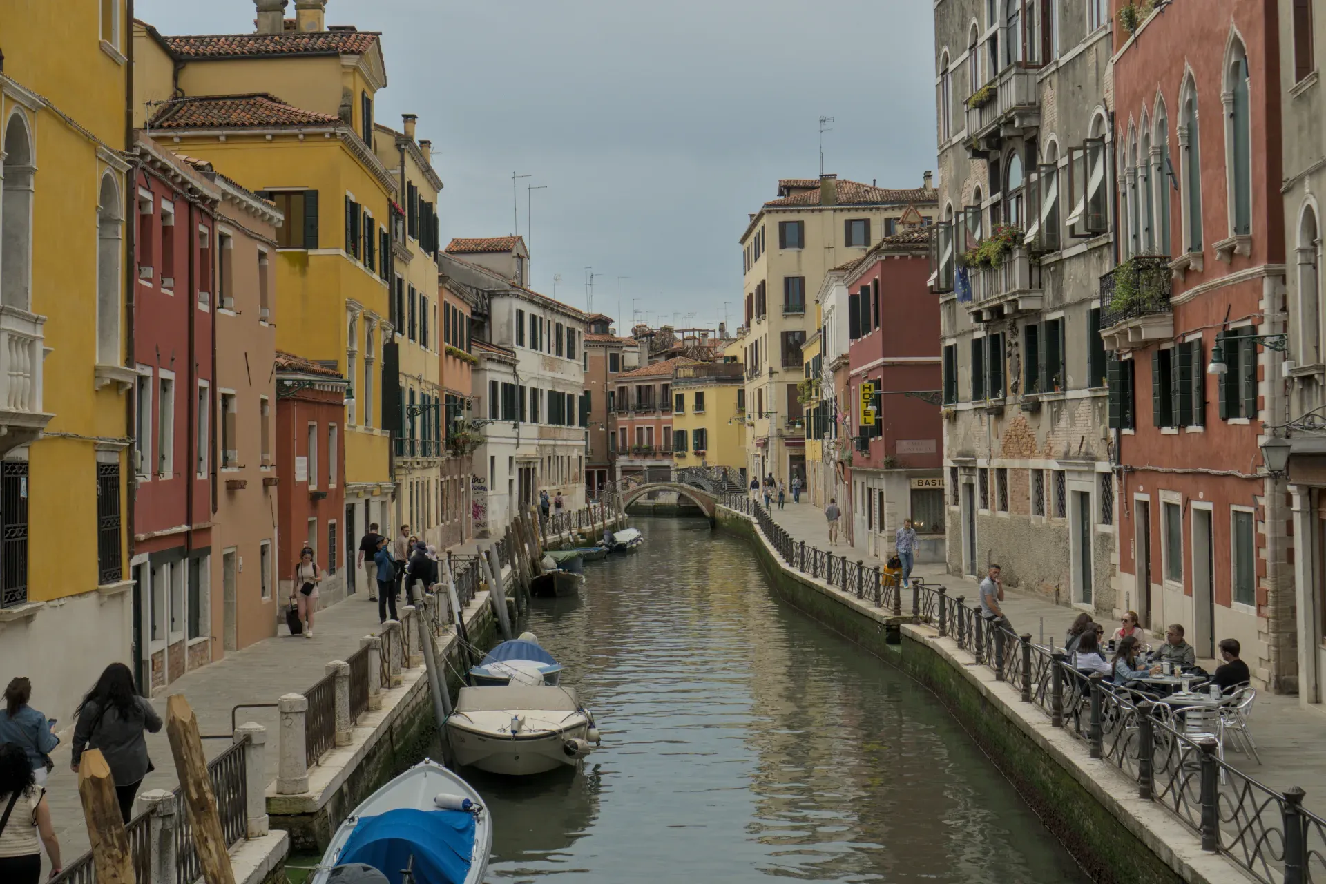 Un callejón estrecho en Venecia con casas coloridas, un canal y barcos, con gente paseando por una acera.