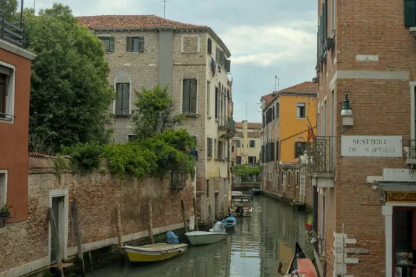 Ein schmaler venezianischer Kanal mit traditionellen Gebäuden, grüner Vegetation und mehreren Booten, die im Wasser treiben.