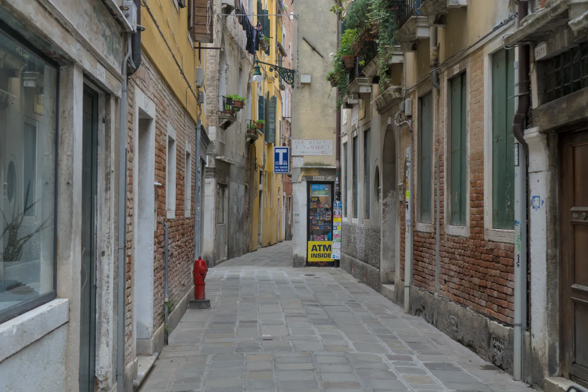 Fotografia di uno stretto vicolo veneziano con acciottolato, muri in mattoni ed edifici storici.