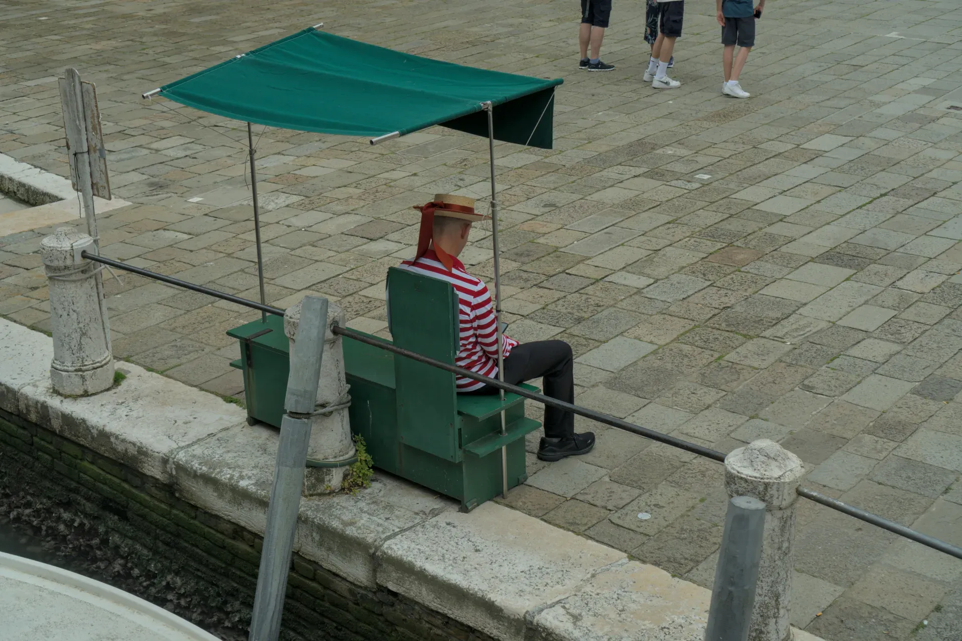 Un gondolier attend à l'ombre sous un auvent vert, vêtu d'une chemise à rayures et d'un chapeau.