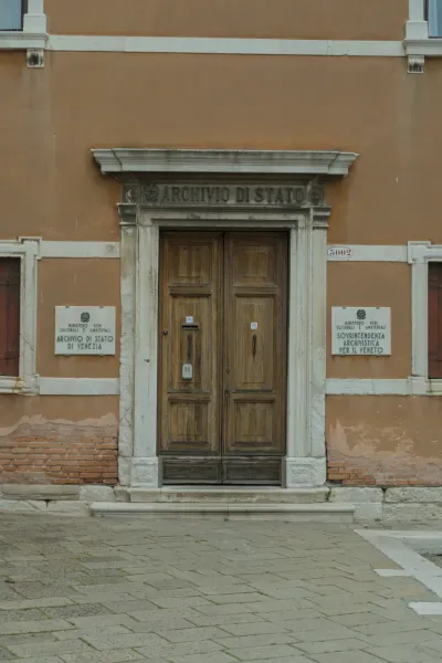 Široka, stara drvena vrata na ulazu u Archivio di Stato u Veneciji, uokvirena kamenim stubovima i oker fasadom.