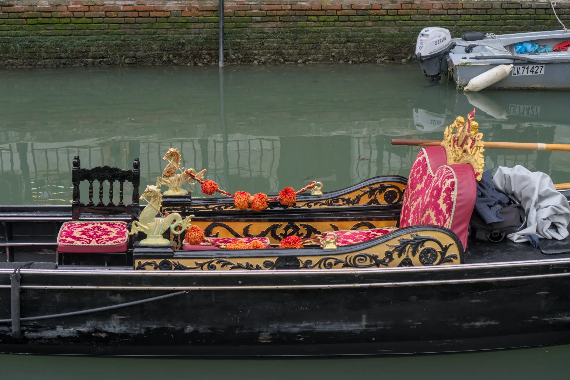 Une gondole vénitienne ornée de décorations rouges et dorées, dérivant sur une eau verte calme contre un mur de briques.