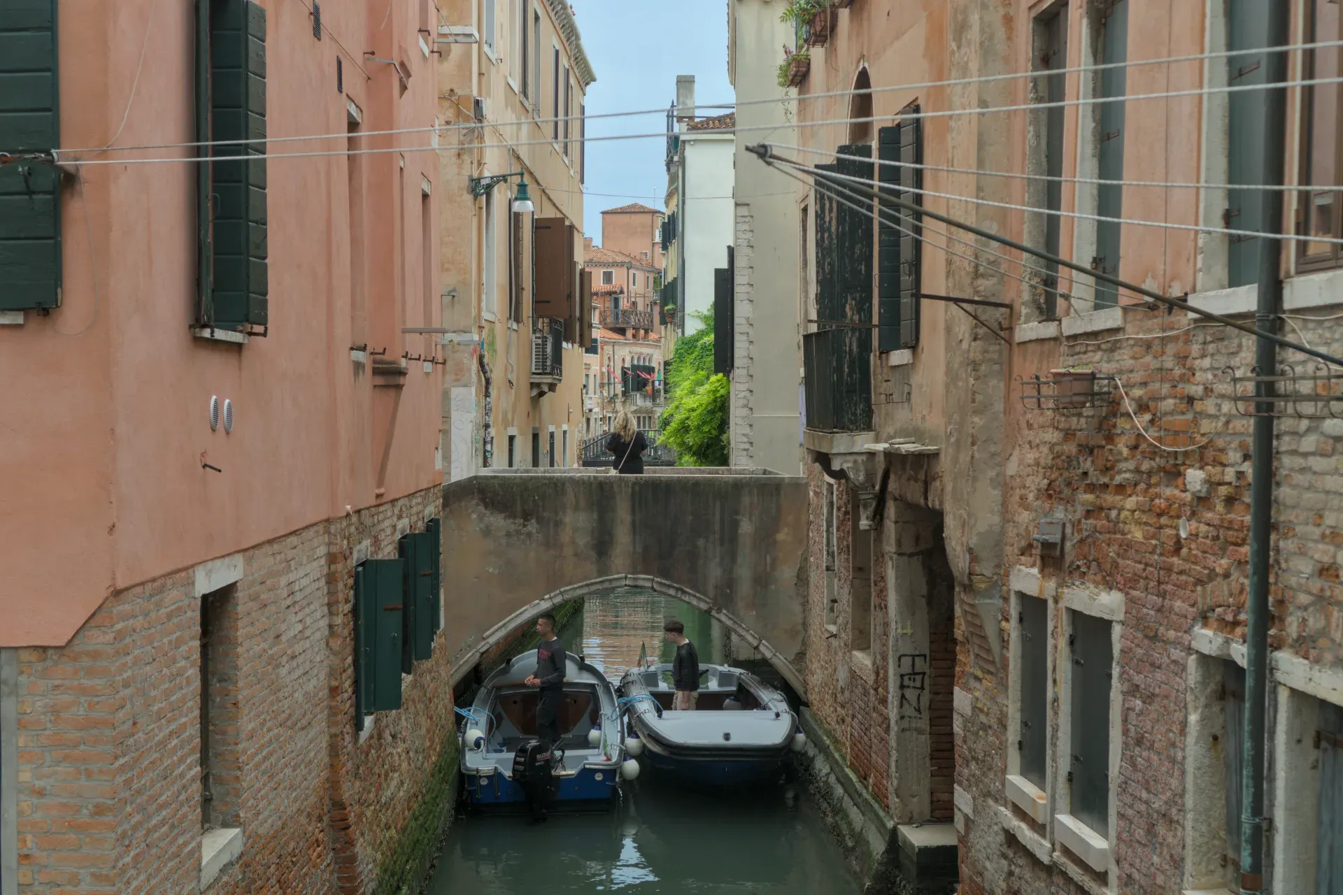Canale veneziano con due barche e persone sotto un ponte, circondato da vecchi edifici in mattoni.