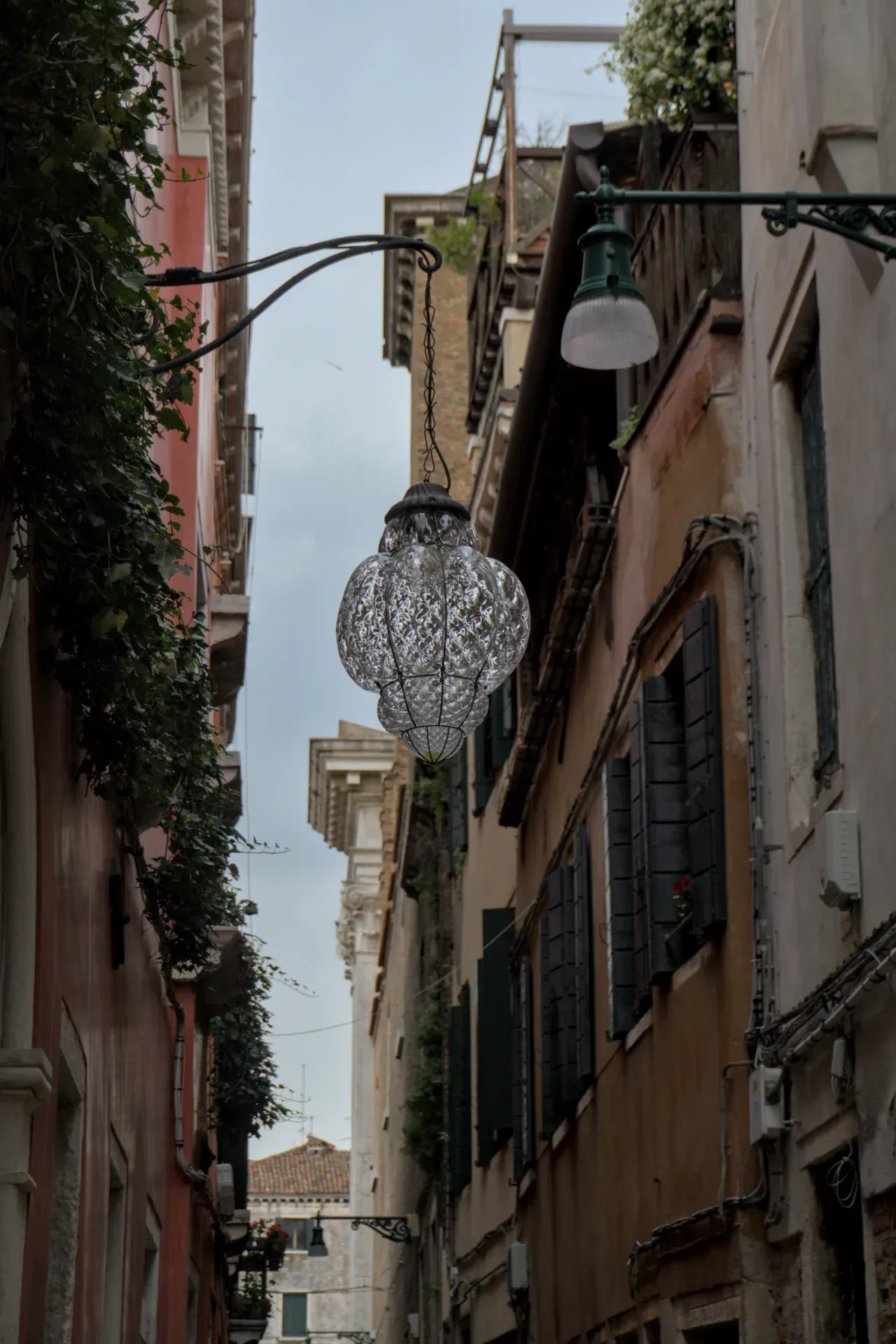 Eine kunstvolle, facettierte Glaslaterne hängt an einer Kette in einer engen Gasse Venedigs, mit Gebäuden und bewachsenen Mauern im Hintergrund.