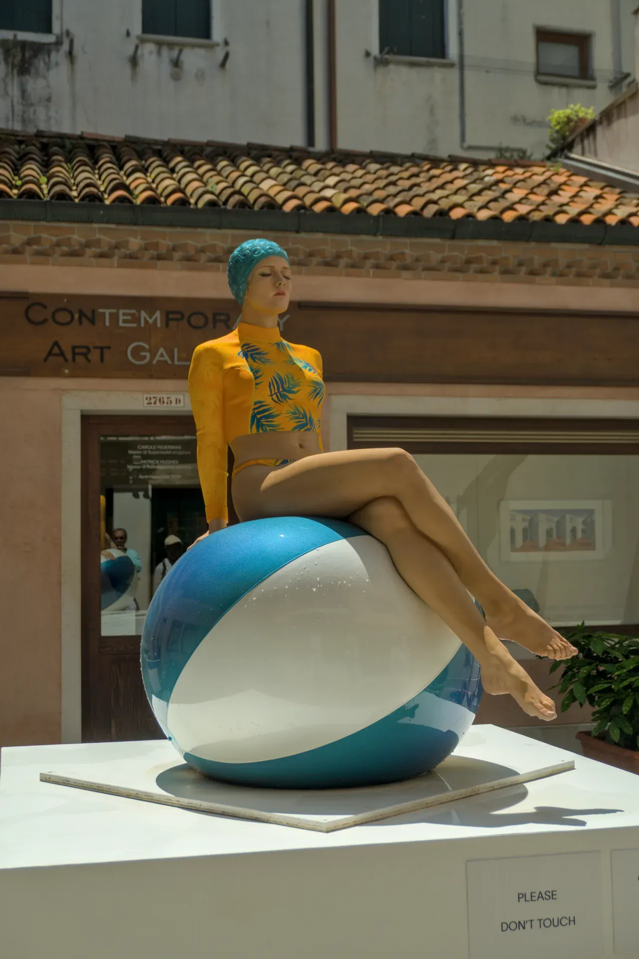 Portrait d'une sculpture de femme assise sur un ballon de plage bleu et blanc à Venise, portant un maillot de bain jaune.