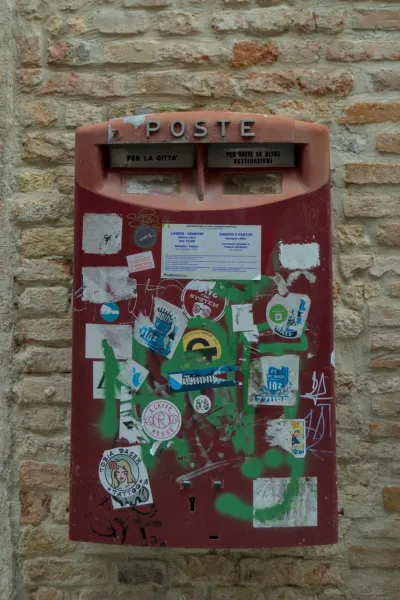 Ein alter, mit Aufklebern und Graffiti verzierter roter Postkasten vor einer Ziegelmauer in Venedig.
