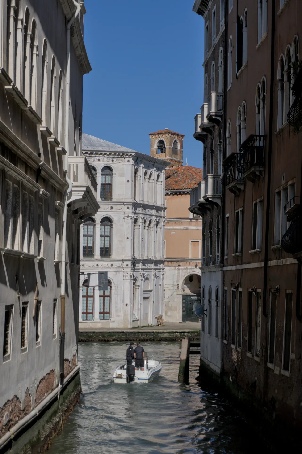 Ein Boot fährt auf einem schmalen venezianischen Kanal, flankiert von historischen Gebäuden, mit einem Glockenturm im Hintergrund.