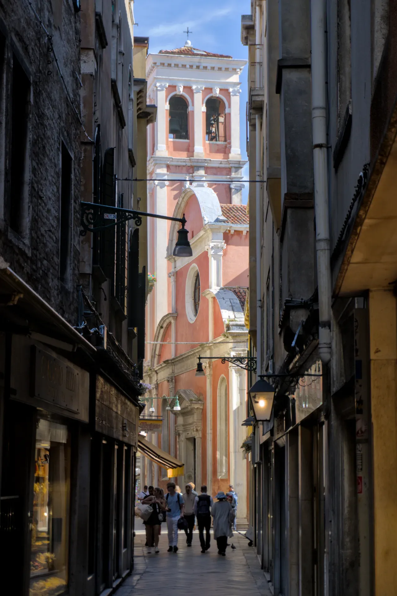 Photographie d'une ruelle étroite à Venise menant à un clocher rose avec des cloches et des arches, avec des gens qui passent.