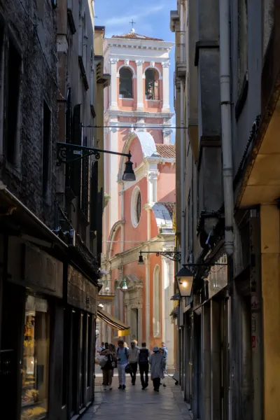 Fotografía de un callejón estrecho en Venecia que conduce a un campanario rosa con campanas y arcos, con gente caminando.