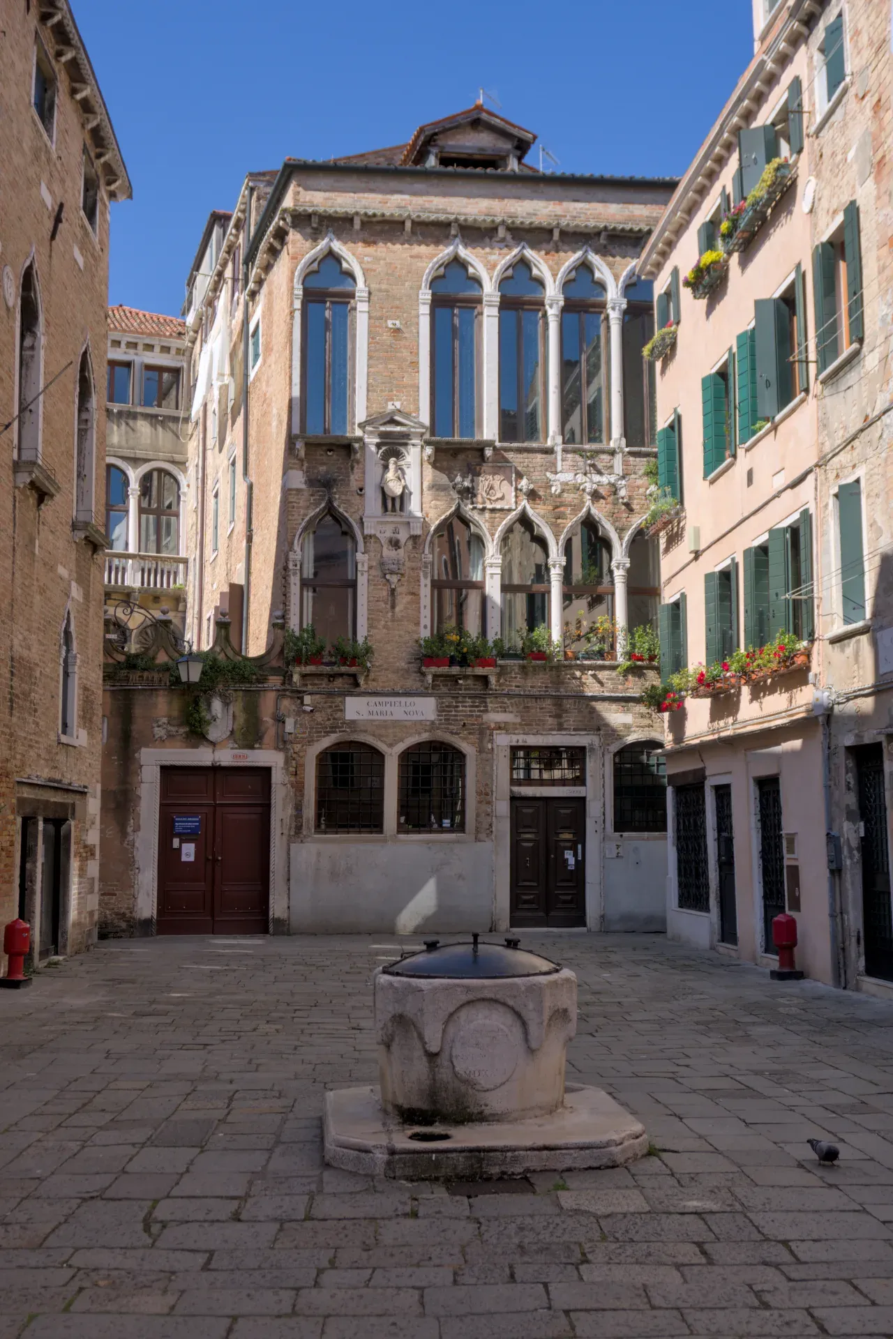 Un patio veneciano soleado con un pozo de piedra en primer plano y edificios históricos con arcos.