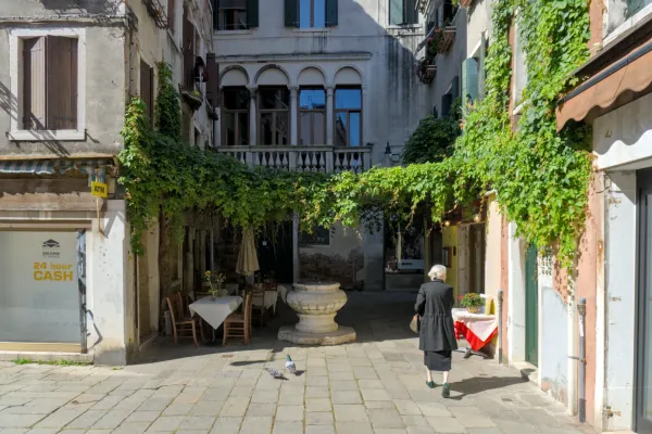 Une femme traverse une ruelle vénitienne verte et envahie par le lierre, avec des tables extérieures et un puits ancien.