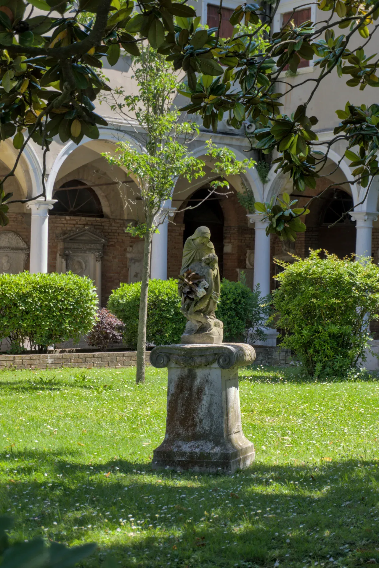 Sculpture en pierre sur un piédestal, entourée d'un jardin verdoyant sur l'île-cimetière de San Michele à Venise.