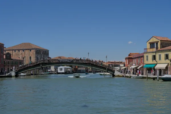 Un puente peatonal de madera en la isla de Murano en Venecia, cruzado por personas, con barcos en el agua y edificios históricos.