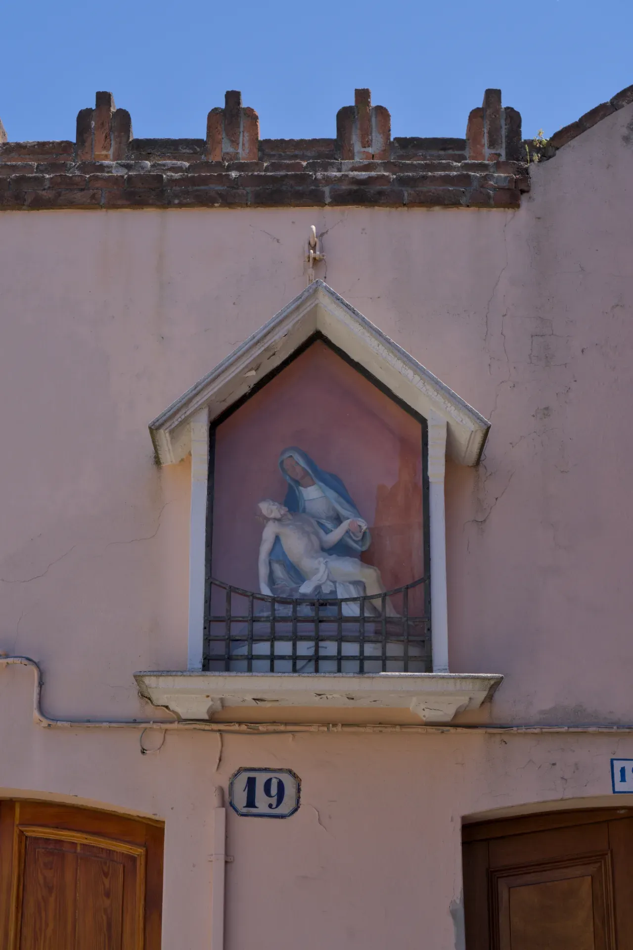 Fine art photograph of a Pietà depiction on the island of Murano, Italy, featuring a pink plastered building and blue sky.