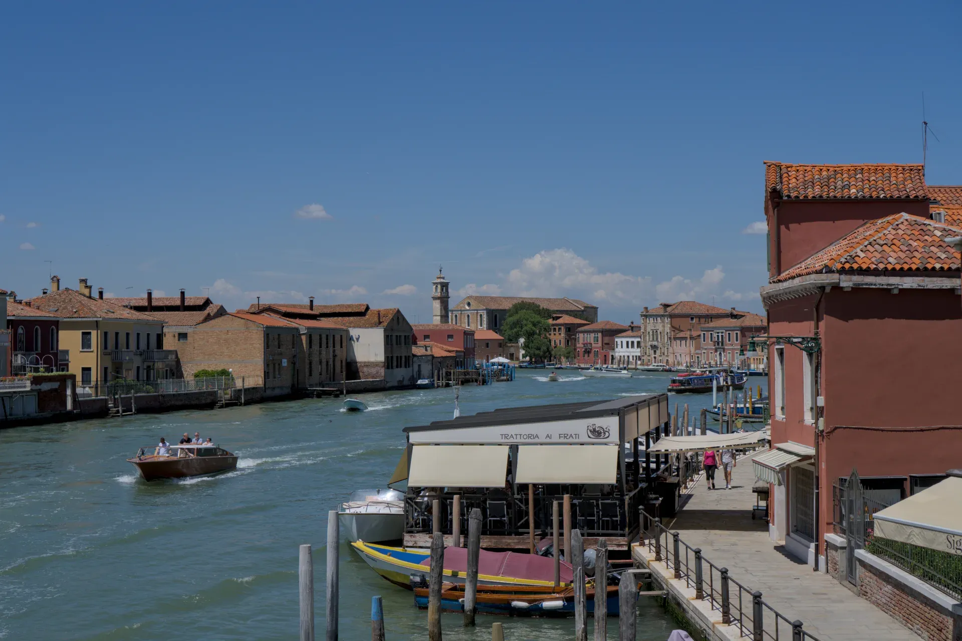 Una vista sulla sponda del canale dell'isola di Murano con acqua, barche e caratteristiche case veneziane sotto un cielo blu.