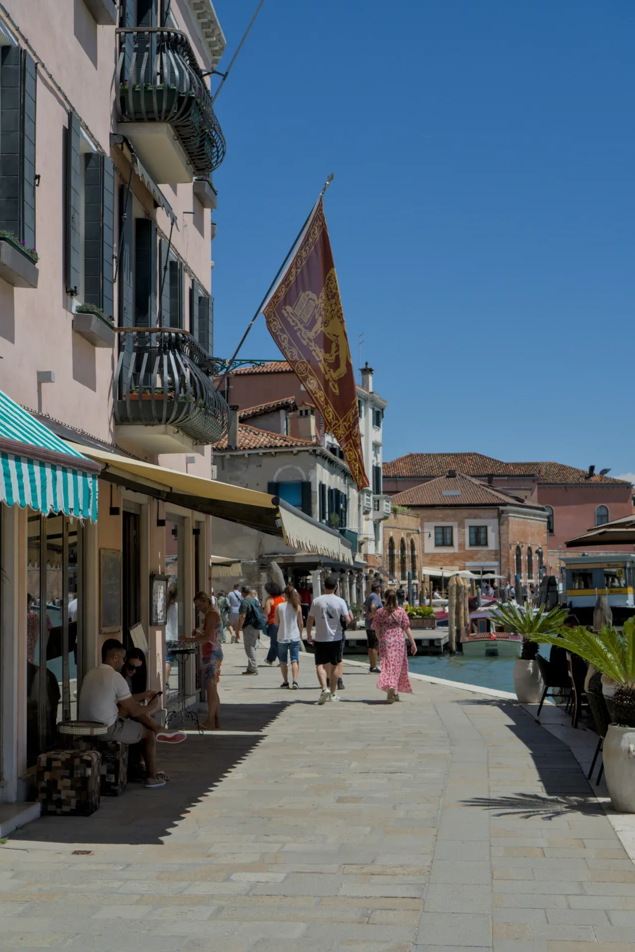 Menschen schlendern entlang eines Kanals auf der Insel Murano, Venedig, unter einer wehenden venezianischen Flagge.