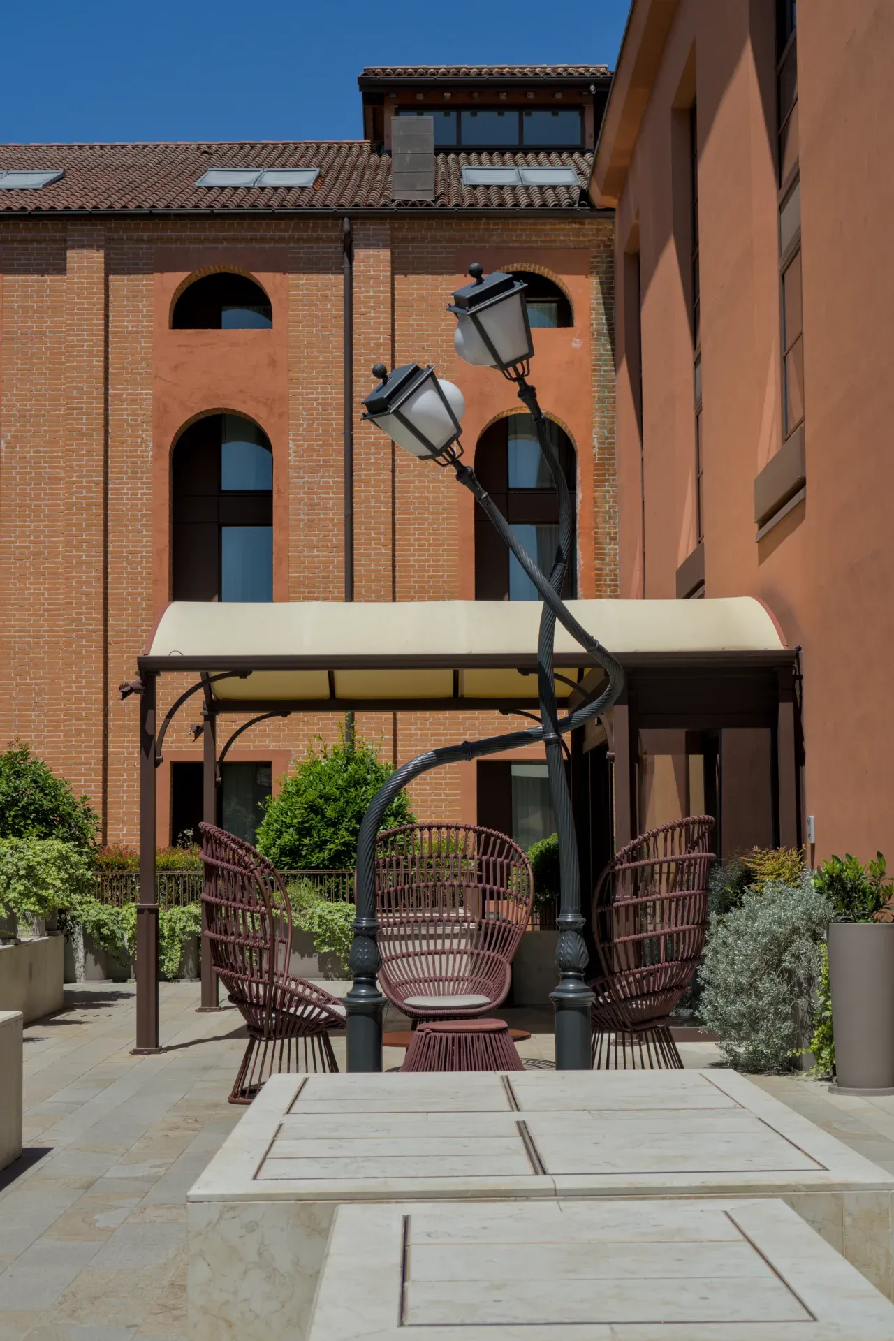 Une terrasse ensoleillée à Murano avec des chaises en osier et un lampadaire élégant devant des bâtiments de briques et de terre cuite.