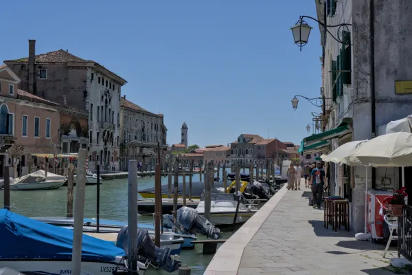 Una vista di un canale a Venezia con barche ormeggiate al molo e case tradizionali sullo sfondo, con Murano in lontananza