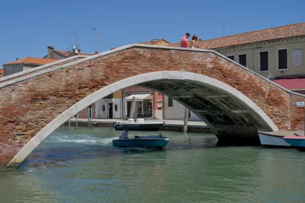 A stone bridge with brick accents spans a canal on the island of Murano, Italy, with boats under the bridge and a couple on the parapet.