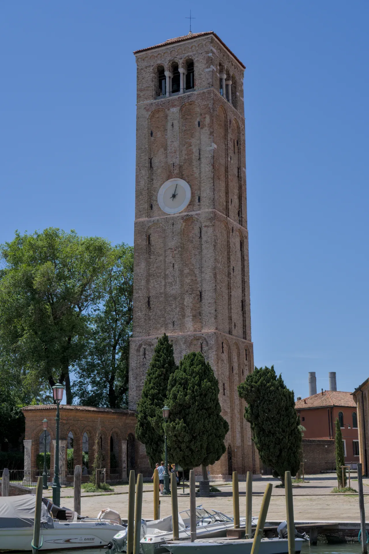 Der Backsteinglockenturm der Insel Murano unter blauem Himmel, mit Bäumen und Wasser davor.
