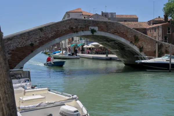 Ponte storico in mattoni sull'isola di Murano, Venezia, che attraversa un canale con piccole barche e caffè.