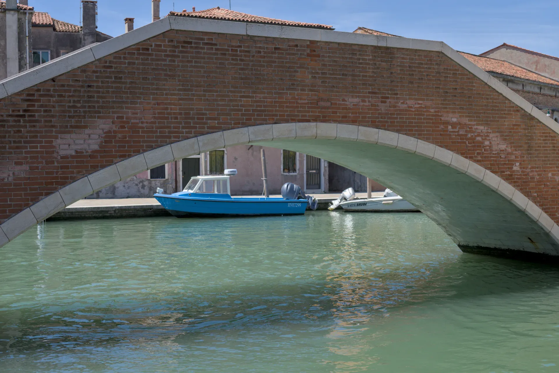 Eine rote Ziegelbrücke mit Steinverzierungen überspannt einen Kanal, auf dem ein blaues Boot liegt, nahe der Insel Murano.