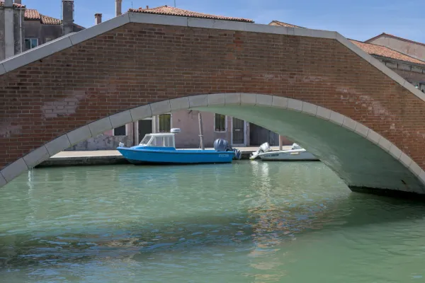 Un ponte in mattoni rossi con decorazioni in pietra sovrasta un canale, con una barca blu adagiata su di esso, vicino all'isola di Murano.