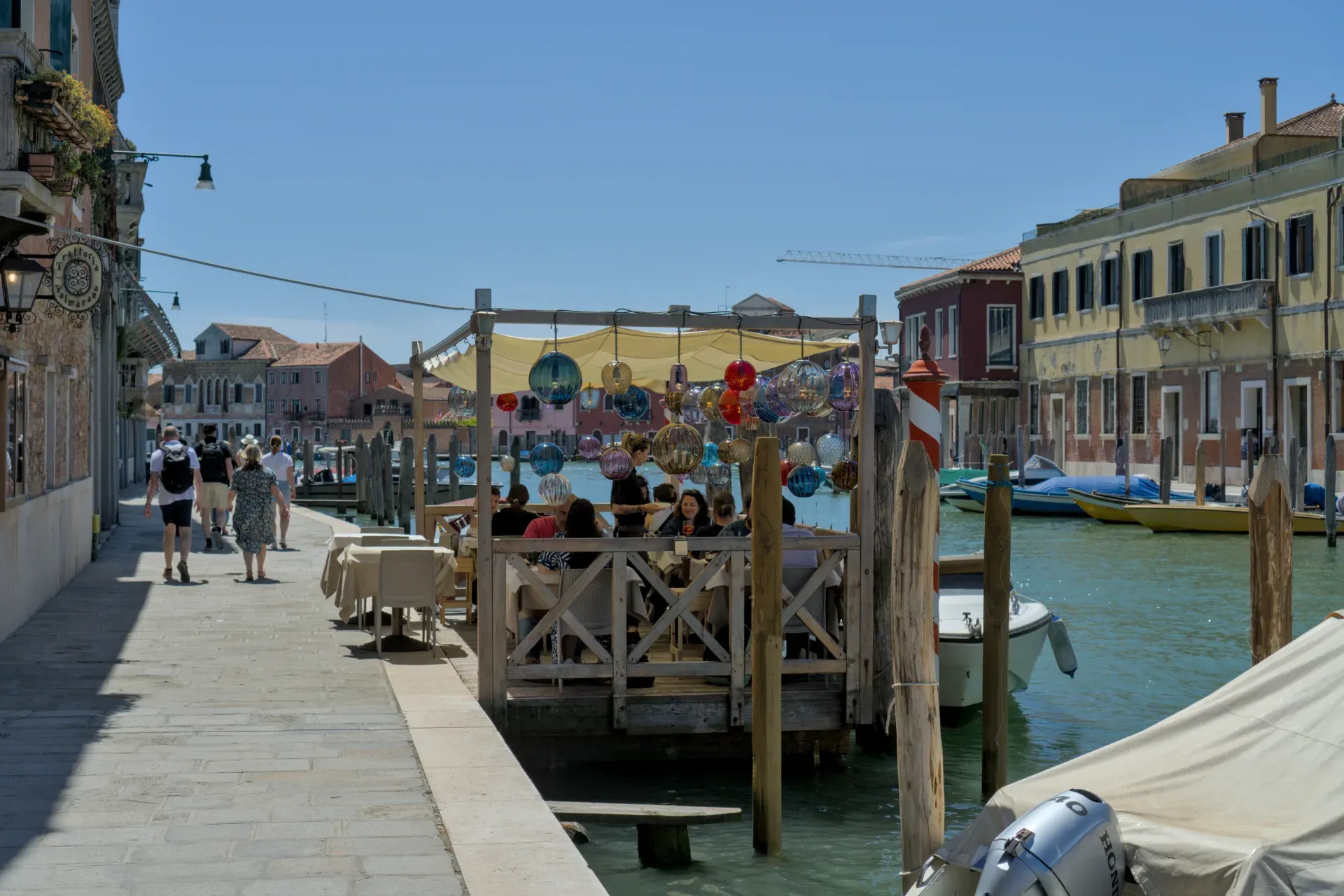 Un canal à Murano avec un restaurant et des lumières en verre colorées, des bâtiments traditionnels et des bateaux en arrière-plan.