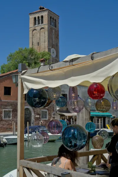 Ručno rađena murano staklena svetla vise iznad kanala u Veneciji sa kulom u pozadini.