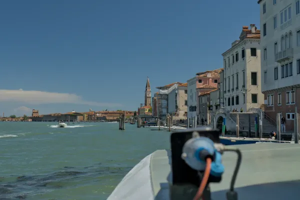 Une journée ensoleillée à Venise, vue sur la rive du canal avec des bâtiments traditionnels se reflétant dans l'eau turquoise et un bateau qui passe.