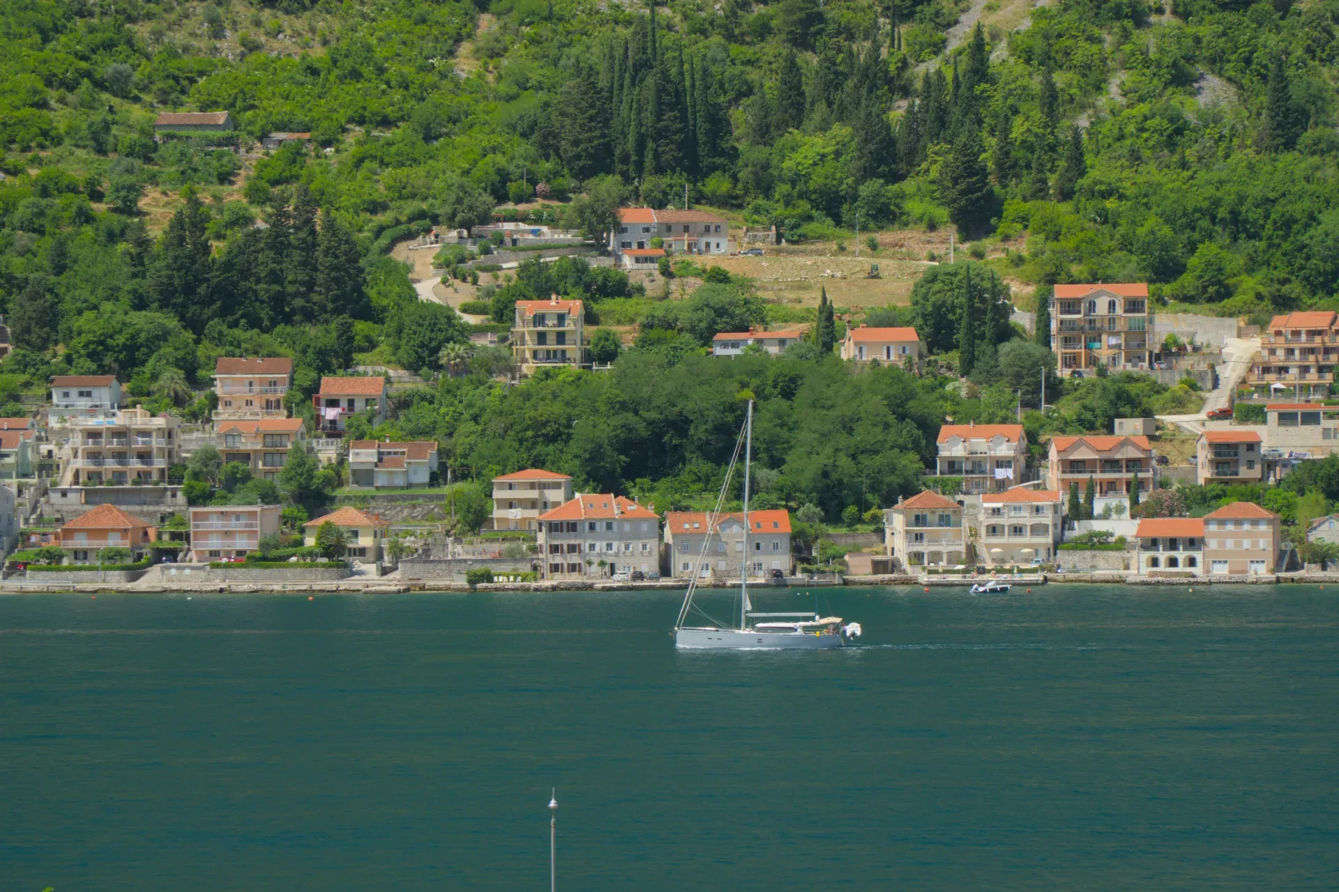 Ein Segelboot auf dem ruhigen Wasser vor einer Küstenstadt mit roten Dächern, die an einem grünen Hügel liegt.